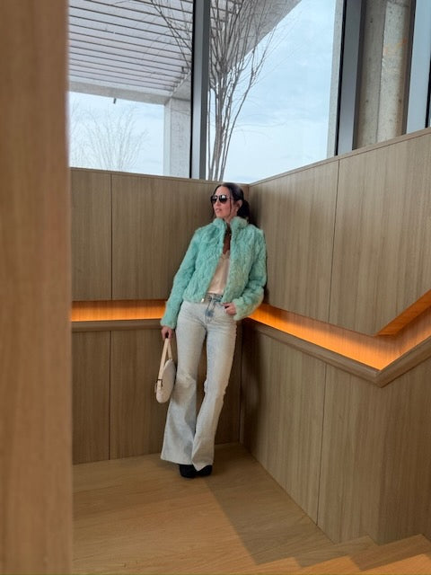 Woman in a light blue jacket and white pants standing in a modern indoor setting with wooden walls and orange lighting.