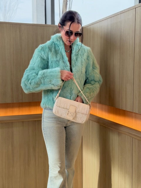 Person wearing a light green fuzzy jacket and holding a beige handbag indoors.