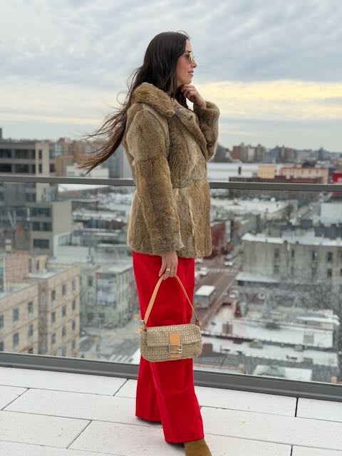 Woman in a fur coat and red pants standing on a rooftop with a cityscape background