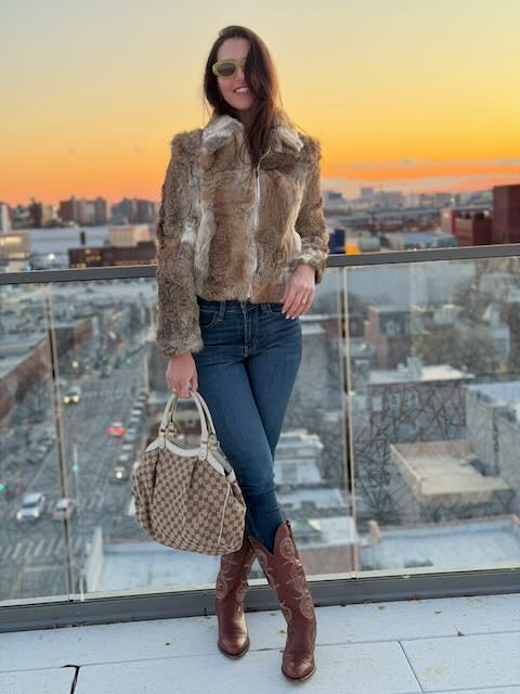 Woman in fur coat and brown boots standing on a rooftop with a cityscape background during sunset.