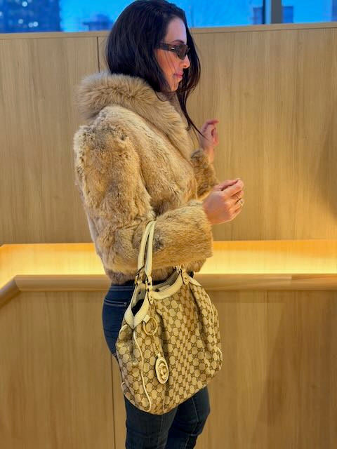 Woman wearing a fur coat and holding a patterned handbag against a wooden wall.