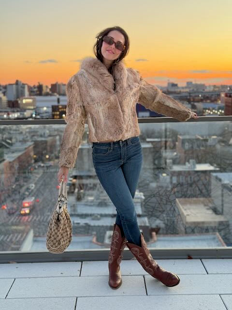 Woman in a fur-lined coat and brown boots standing on a rooftop with a cityscape and sunset in the background.